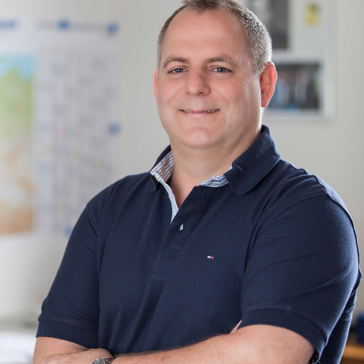 Ein Mann mit kurzem Haar und einem blauen Poloshirt steht lächelnd im Büro mit verschränkten Armen.